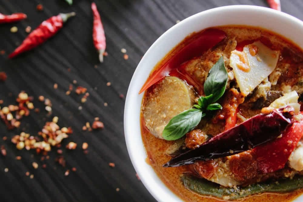 A vibrant bowl of spicy curry garnished with basil and topped with red chilies, showcasing fresh vegetables.