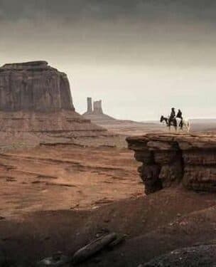 Two horseback riders on a rocky plateau with towering cliffs in the background.
