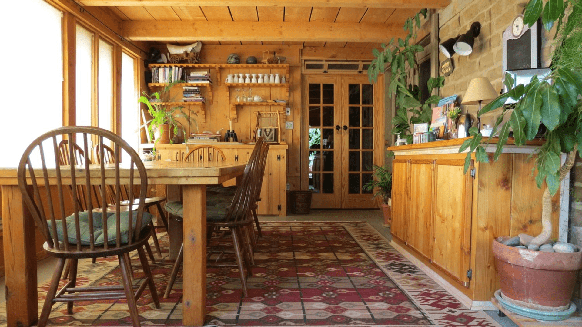A cozy wooden dining area with a table, chairs, and plants, featuring large windows and warm light.