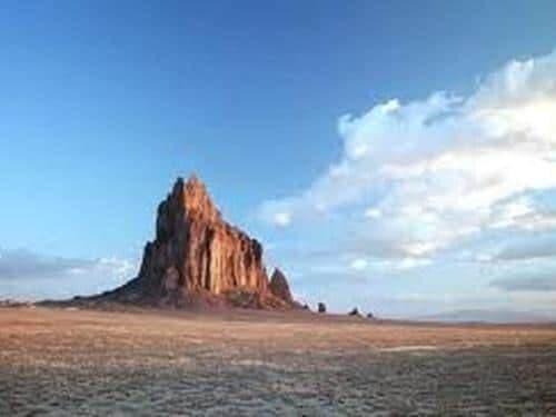 A towering rock formation rises from a barren landscape under a clear blue sky.
