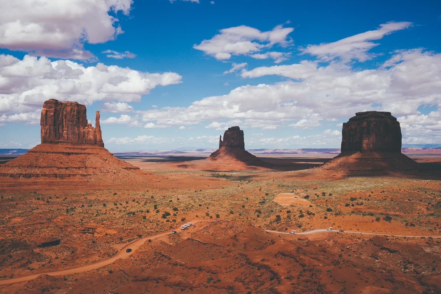 A sweeping view of red desert landscape featuring three towering rock formations under a blue sky with clouds.