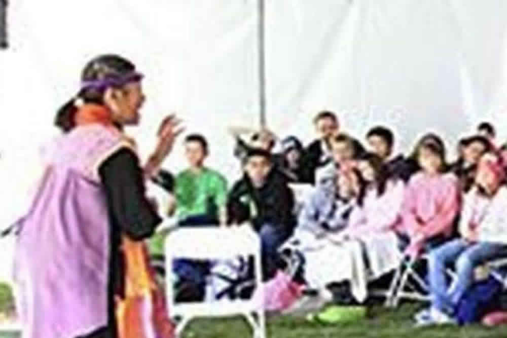 A performer gestures while speaking to an audience seated in folding chairs under a tent.
