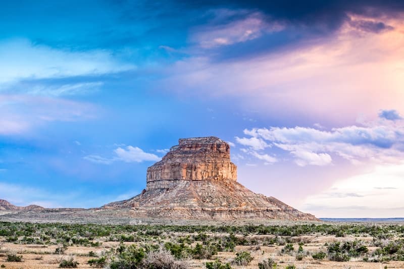 A dramatic rock formation rises above a vast, arid landscape under a colorful sky.