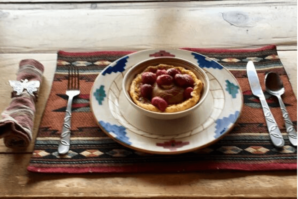 A dessert with raspberries sits on a decorative plate placed on a patterned tablecloth, accompanied by cutlery.